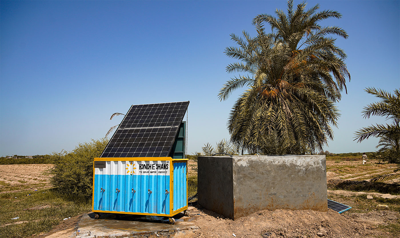 Our technology OASIS Box in a rural landscape, with a bright blue sky above. To the right, a mature palm tree stands tall, and in the background, there are fields with sparse vegetation, emphasizing the arid environment. A concrete well is adjacent to the container.